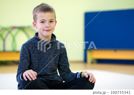 Young child boy sitting and relaxiong on the floor inside sports room in a school after training. 100735341