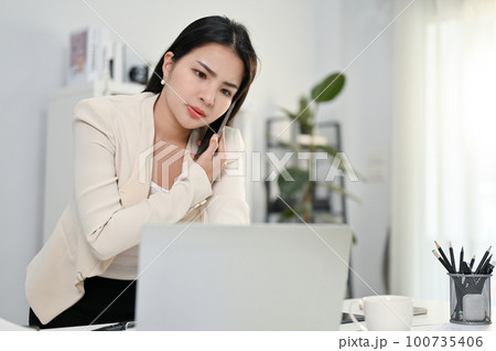 Stressed and serious Asian female boss talking on the phone with her employee 100735406