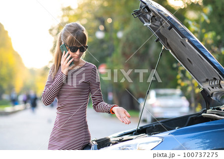 Upset young woman driver talking angrily on cell phone with assistance service near a broken car with open hood on a city street. Upset young woman driver talking angrily on cell phone with assistance service near a broken car with open hood on a city street. 100737275