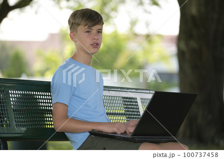 Teenager school boy typing on laptop computer sitting on bench outdoors on summer sunny day. Education during quarantine concept Teenager school boy typing on laptop computer sitting on bench outdoors on summer sunny day. Education during quarantine concept 100737458