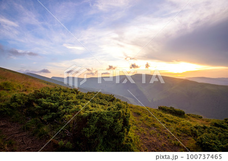 Summer evening mountain landscape with grassy hills and distant peaks at colorful sunset. 100737465