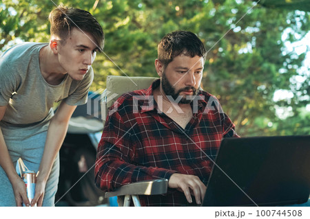 Caucasian bearded man and teenage son working wing with laptop outdoors. Camping concept 100744508