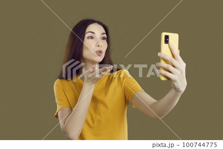 Pretty young woman holding her modern cellphone, blowing air kiss and taking selfie 100744775