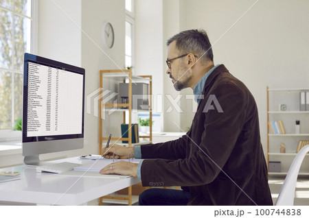 Portrait of serious, attentive, mature businessman man with glasses, sitting at his workplace and looking at his computer screen. 100744838