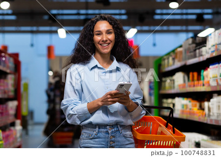 Shopping in a supermarket. A young Latin American woman stands between rows of shelves with a cart. He holds the phone, has a list, writes messages, orders and pays online. Smiling at the camera. 100748831