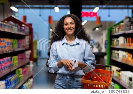 Shopping in a supermarket. A young Hispanic woman walks between rows of shelves with a shopping cart. He holds the phone in his hands, smiles at the camera. Shopping in a supermarket. A young Hispanic woman walks between rows of shelves with a shopping cart. He holds the phone in his hands, smiles at the camera. 100748835