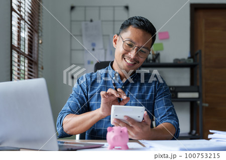 Portrait of an Asian businessman using a calculator to calculate his savings from SME operations, with a pink piggy bank as keep money concept. Portrait of an Asian businessman using a calculator to calculate his savings from SME operations, with a pink piggy bank as keep money concept. 100753215