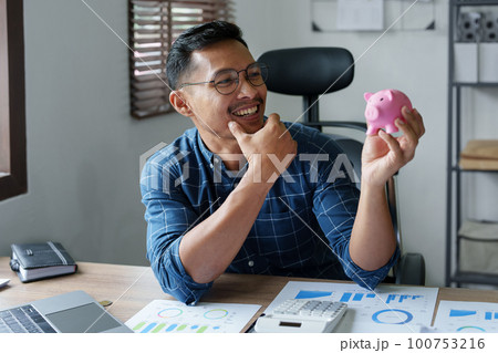 Portrait of an Asian businessman using a calculator to calculate his savings from SME operations, with a pink piggy bank as keep money concept. Portrait of an Asian businessman using a calculator to calculate his savings from SME operations, with a pink piggy bank as keep money concept. 100753216