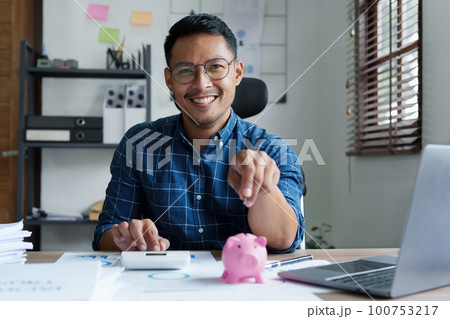 Portrait of an Asian businessman using a calculator to calculate his savings from SME operations, with a pink piggy bank as keep money concept. Portrait of an Asian businessman using a calculator to calculate his savings from SME operations, with a pink piggy bank as keep money concept. 100753217