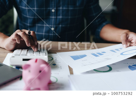 Portrait of an Asian businessman using a calculator to calculate his savings from SME operations, with a pink piggy bank as keep money concept. Portrait of an Asian businessman using a calculator to calculate his savings from SME operations, with a pink piggy bank as keep money concept. 100753292