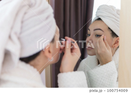 A woman applies black mascara to her eyelashes with a makeup brush. Young beautiful woman applying mascara makeup on her eyes in the bathroom 100754009