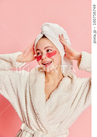 Portrait of smiling happy beautiful girl wearing bathrobe and towel using eye patches for skin refresh over pink background Portrait of smiling happy beautiful girl wearing bathrobe and towel using eye patches for skin refresh over pink background 100756749