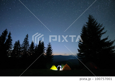 Silhouette of lonely hiker resting besides burning bonfire near illuminated tourist tents on camping site in dark mountains under night sky with stars. Active lifestyle and outdoor living concept. Silhouette of lonely hiker resting besides burning bonfire near illuminated tourist tents on camping site in dark mountains under night sky with stars. Active lifestyle and outdoor living concept. 100757001