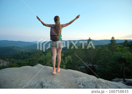 Hiker woman standing on mountain footpath raising up hands enjoying evening nature during travel on wilderness trail. Lonely female traveler traversing high hilltop route. Healthy lifestyle concept Hiker woman standing on mountain footpath raising up hands enjoying evening nature during travel on wilderness trail. Lonely female traveler traversing high hilltop route. Healthy lifestyle concept 100758635