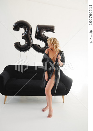 a naked blonde with tattoos in a black coat by a black sofa. birthday 100760511
