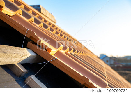 Closeup of metal montage anchor for installation of yellow ceramic roofing tiles mounted on wooden boards covering residential building roof under construction. Closeup of metal montage anchor for installation of yellow ceramic roofing tiles mounted on wooden boards covering residential building roof under construction. 100760723