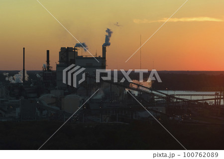Aerial view of wood processing factory with smoke from production process polluting atmosphere at plant manufacturing yard. Industrial site at sunset Aerial view of wood processing factory with smoke from production process polluting atmosphere at plant manufacturing yard. Industrial site at sunset 100762089