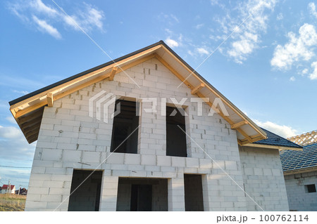 Aerial view of unfinished house with aerated lightweight concrete walls and wooden roof frame covered with metallic tiles under construction. Aerial view of unfinished house with aerated lightweight concrete walls and wooden roof frame covered with metallic tiles under construction. 100762114