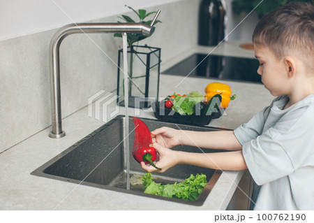 Six-year-old boy washing vegetables in the kitchen sink. Side view. Six-year-old boy washing vegetables in the kitchen sink. Side view. 100762190