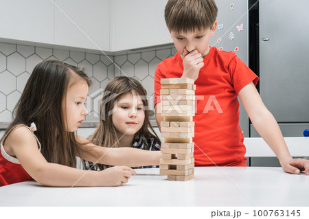 Children of concentrated thoughtful girls, boy play wooden brick tower game on table. Balance challenge, moving blocks 100763145