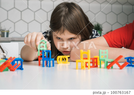 Thoughtful, focused little girl building and holding small green chair toy near many colorful chair toys on white table 100763167
