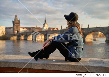 Stylish beautiful young woman wearing black hat sitting on Vltava river shore in Prague with Charles Bridge on background. Elegant retro lady fine art portrait. 100765314