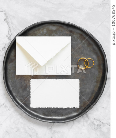 Black plate with a blank card, envelope and wto wedding rings top view, mockup 100768545