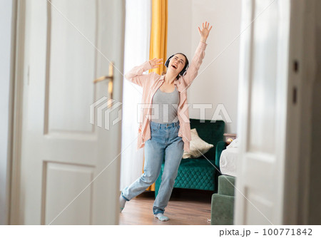 Happy young Caucasian happy woman wearing headphones dance and enjoy life. Window in background. Leisure in home Happy young Caucasian happy woman wearing headphones dance and enjoy life. Window in background. Leisure in home 100771842