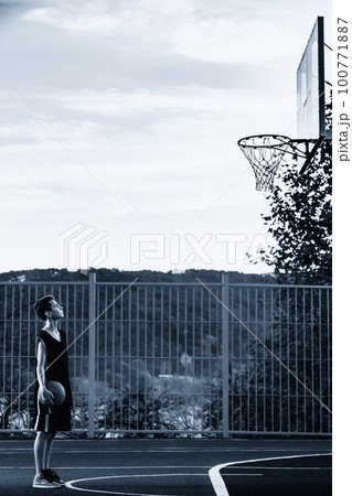 Sports and basketball. A young teenager in a black tracksuit stands on the Playground with a ball in his hands and looks at the basketball Hoop. Vertical. Copy space. Side view. Monochrome Sports and basketball. A young teenager in a black tracksuit stands on the Playground with a ball in his hands and looks at the basketball Hoop. Vertical. Copy space. Side view. Monochrome 100771887