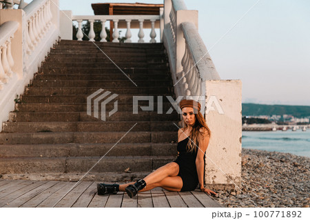 Fashion. Sexy blonde in a black dress and hat posing sitting on the steps. In the distance you can see the sea Fashion. Sexy blonde in a black dress and hat posing sitting on the steps. In the distance you can see the sea 100771892