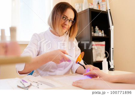 Medicine, nail Polish and prosthetic nail. In the nail salon, a procedure for prosthetics of the nail on the hand is carried out. In the hands of a female master in glasses, ultraviolet lamp 100771896