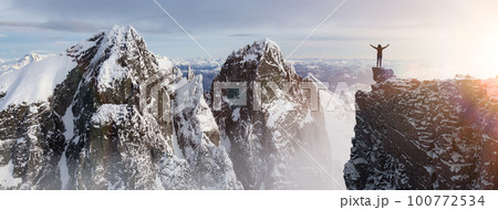 Adventurous Woman Hiker standing on top of icy peak with rocky mountains in background. 100772534