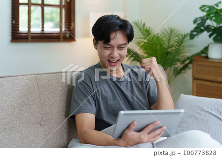 Portrait of an Asian teenager using a digital tablet on the sofa while relaxing at home. 100773828