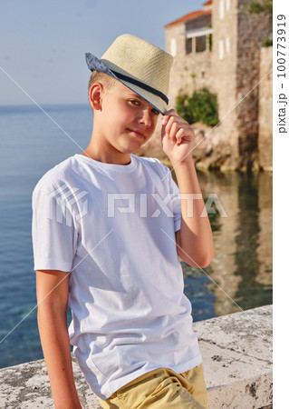 Portrait of a teenager boy in a white t-shirt and straw hat sitting near the sea. Portrait of a teenager boy in a white t-shirt and straw hat sitting near the sea. 100773919