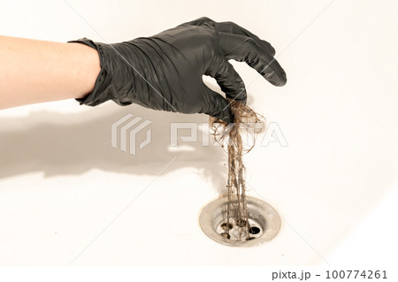 A bathroom drain clogged with dirty hair and slime. A female hand in a black latex glove pulls a long messy tuft of hair out of the drain 100774261