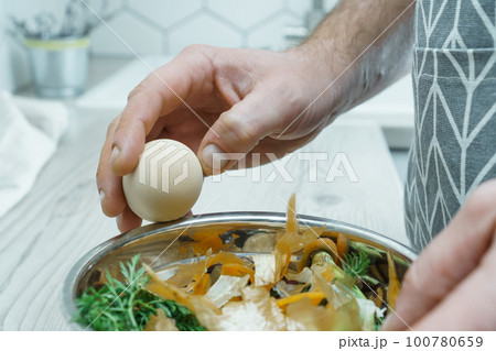 Male hands peeling shell of boiled egg over bowl of vegetable peelings closeup. Cook on kitchen table, food scrap. 100780659