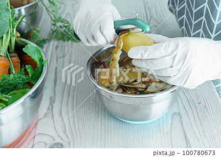 Human hands in white domestic gloves peeling potato with peeler over bowl of peelings. Food scrap, zero waste. Human hands in white domestic gloves peeling potato with peeler over bowl of peelings. Food scrap, zero waste. 100780673