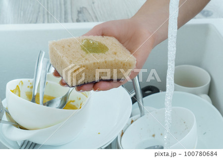 Human hand wash white dinnerware with dish soap on sponge under tap water. Pile of kitchen utensils in sink closeup. Human hand wash white dinnerware with dish soap on sponge under tap water. Pile of kitchen utensils in sink closeup. 100780684