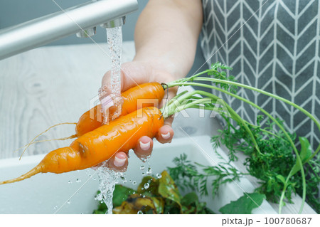 Female hands washing fresh carrot under water jet in kitchen sink closeup. Dirty raw orange carrot with green haulm. 100780687