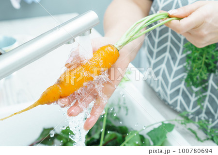 Female hands washing fresh carrot under tap water jet in kitchen sink closeup. Dirty raw orange carrot with green haulm. Female hands washing fresh carrot under tap water jet in kitchen sink closeup. Dirty raw orange carrot with green haulm. 100780694