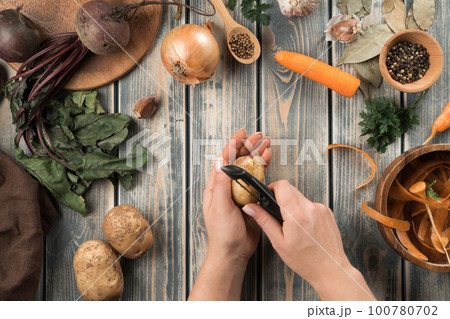 Human hands peel potatoes with vegetable peeler, flat lay. Mix of fresh vegetables on wooden table. Zero waste concept. Human hands peel potatoes with vegetable peeler, flat lay. Mix of fresh vegetables on wooden table. Zero waste concept. 100780702