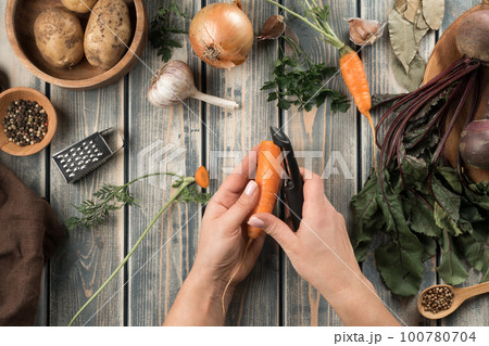 Human hands peel ripe carrot with vegetable peeler, flat lay. Fresh vegetables on wooden table. Zero waste concept. Human hands peel ripe carrot with vegetable peeler, flat lay. Fresh vegetables on wooden table. Zero waste concept. 100780704