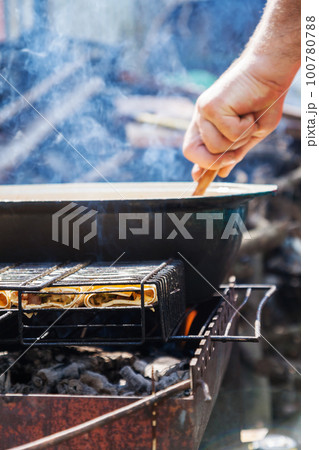 On the grill is a cauldron in which to cook meat and vegetables. The man's hand prepares food 100780788