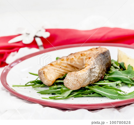 Baked steak trout fish on green leaves of rucola lettuce, lemon, served on white plate serving, top view Baked steak trout fish on green leaves of rucola lettuce, lemon, served on white plate serving, top view 100780828