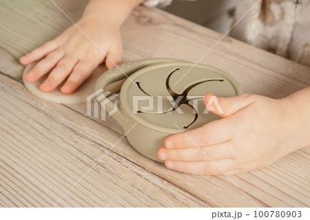 Unrecognizable child holding pastel gray silicone snack cup near lid at wooden table. Baby accessories, tableware 100780903
