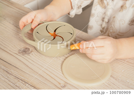 Hands of child take piece of carrot out from pastel gray silicone snack cup at wooden table. Baby accessories, tableware 100780909