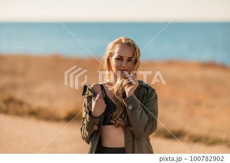 Portrait blonde sea cape. A calm young blonde in an unbuttoned khaki raincoat stands on the seashore, under the raincoat there is a black skirt and top Portrait blonde sea cape. A calm young blonde in an unbuttoned khaki raincoat stands on the seashore, under the raincoat there is a black skirt and top 100782902