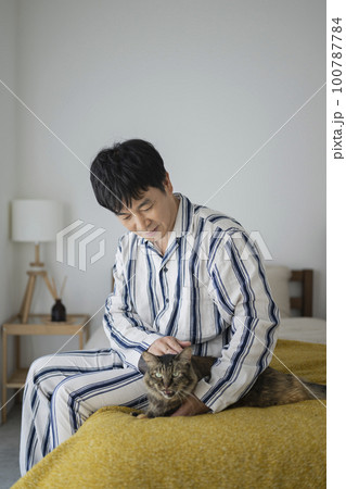 ベッドに座って猫を撫でるパジャマ姿の50代男性 100787784