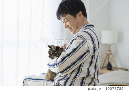 ベッドに座って猫を抱くパジャマ姿の50代男性 100787894