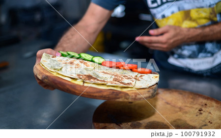 A man presents a wooden plate with a tortilla, cucumber, and tomatoes in the kitchen. 100791932